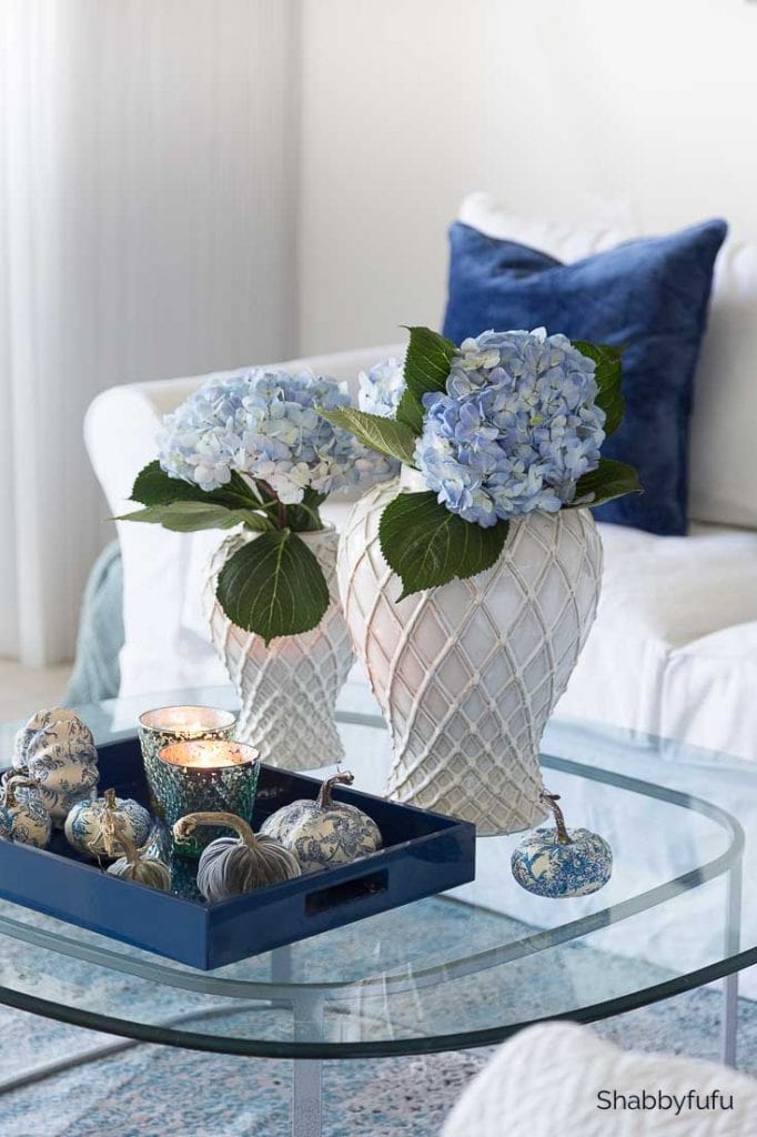 Glass table with blue flowers in white vases and mini pumpkins on table.